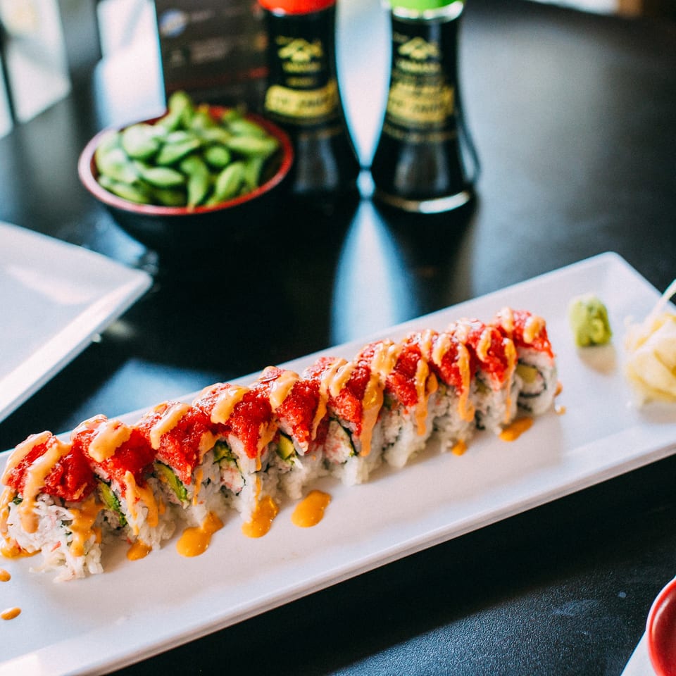 Sushi Omakase's long sushi roll with red topping and orange sauce, with edamame and soy sauce.