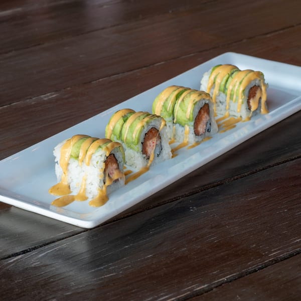 Four sushi rolls with avocado slices and a creamy orange sauce on a white rectangular plate.